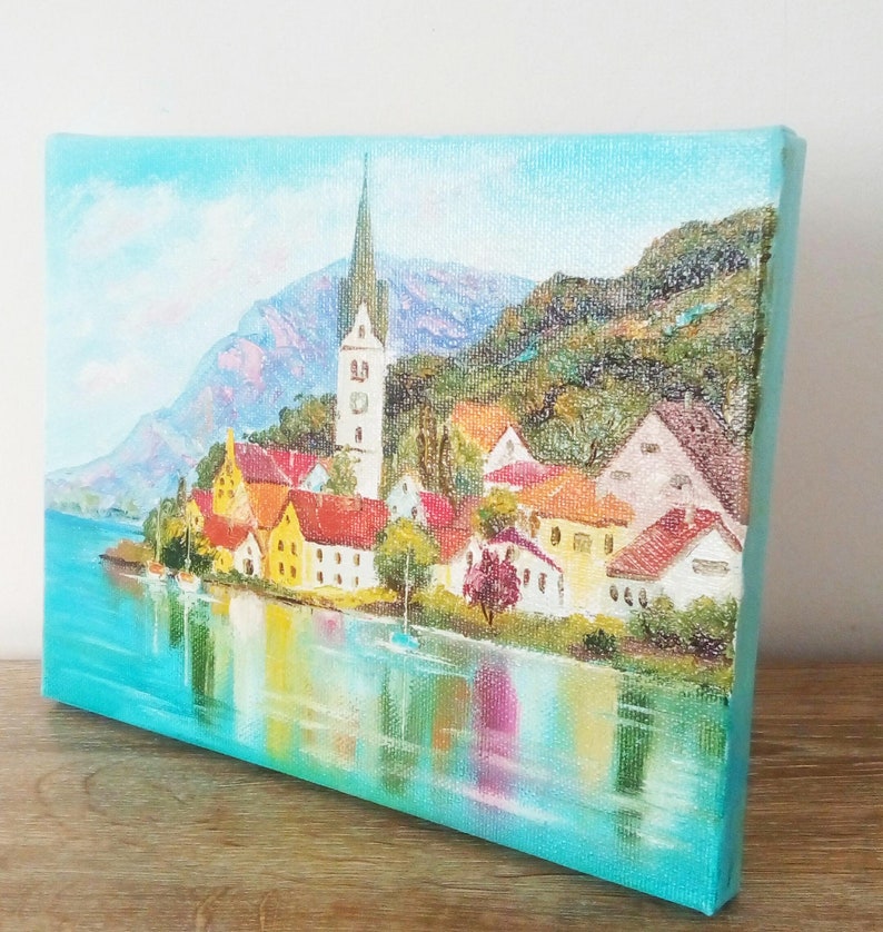 May include: A painting depicting a quaint village with a church spire, nestled in a valley with mountains in the background. The village is located on a lake with colourful reflections of the buildings in the water.