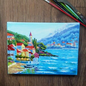 May include: A painting depicting a small town nestled on a hillside overlooking a blue expanse of water. The town features colourful buildings with red roofs and verdant trees. The water is a vibrant blue with whitecaps and the sky is a light blue.