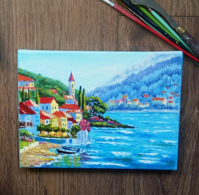 May include: A painting depicting a small town nestled on a hillside overlooking a blue expanse of water. The town features colourful buildings with red roofs and verdant trees. The water is a vibrant blue with whitecaps and the sky is a light blue.