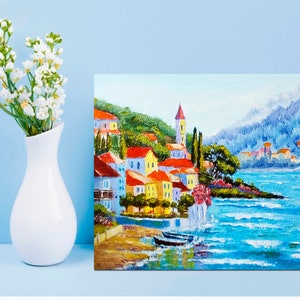 May include: A colourful painting of a seaside town with red-roofed buildings, a church steeple, and a blue ocean with whitecaps. The town is nestled on a hillside with green trees and a mountain in the background.