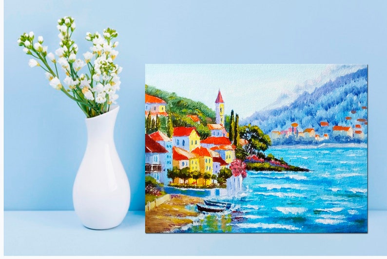 May include: A colourful painting of a seaside town with red-roofed buildings, a church steeple, and a blue ocean with whitecaps. The town is nestled on a hillside with green trees and a mountain in the background.