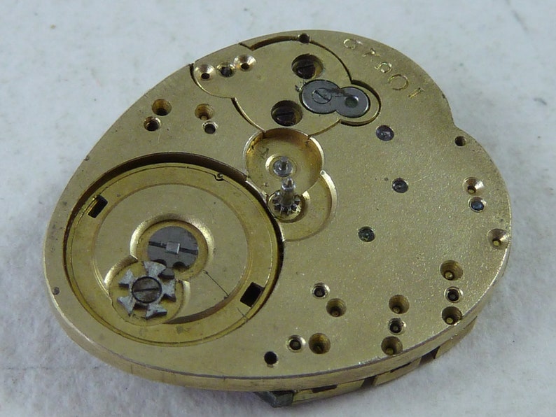 Puede incluir: Un movimiento de reloj de color dorado con engranajes intrincados y una base en forma de coraz&oacute;n. El movimiento est&aacute; etiquetado como "10501".