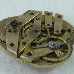Puede incluir: Primer plano de un movimiento de reloj antiguo desmontado con engranajes de lat&oacute;n y una aguja de plata. El movimiento del reloj est&aacute; dispuesto sobre una superficie blanca.