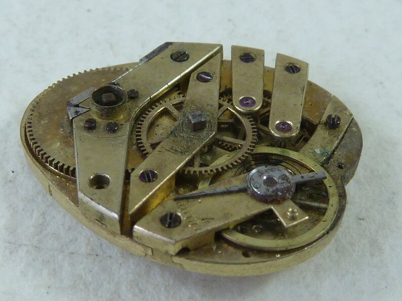 Puede incluir: Primer plano de un movimiento de reloj antiguo desmontado con engranajes de lat&oacute;n y una aguja de plata. El movimiento del reloj est&aacute; dispuesto sobre una superficie blanca.