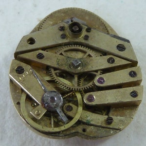 Puede incluir: Primer plano de un movimiento de reloj de tono dorado con engranajes intrincados y un peque&ntilde;o componente redondo de color plateado. El movimiento se coloca sobre un fondo blanco.