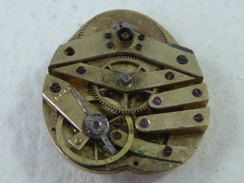 Puede incluir: Primer plano de un movimiento de reloj de tono dorado con engranajes intrincados y un peque&ntilde;o componente redondo de color plateado. El movimiento se coloca sobre un fondo blanco.
