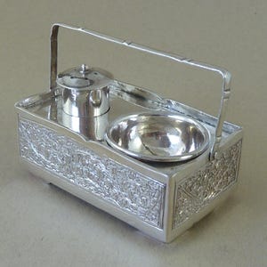 May include: A silver salt and pepper shaker set with a handle. The set is decorated with an intricate floral pattern and has a small bowl for salt and a small jar with a lid for pepper.