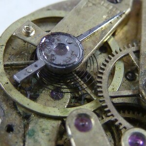 Puede incluir: Primer plano de un mecanismo de reloj vintage con una caja de metal dorado, un v&aacute;stago de cuerda plateado y un muelle negro. El mecanismo es intrincado y detallado, con engranajes y otros componentes visibles.