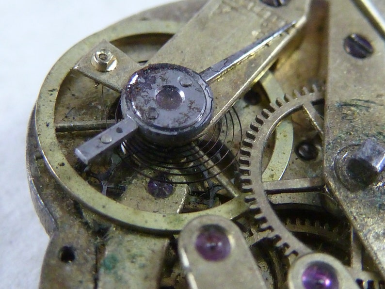 Puede incluir: Primer plano de un mecanismo de reloj vintage con una caja de metal dorado, un v&aacute;stago de cuerda plateado y un muelle negro. El mecanismo es intrincado y detallado, con engranajes y otros componentes visibles.