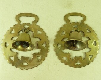 May include: Two antique brass horse brasses with a bell hanging from each. The brasses are circular with a decorative design of hearts and a cutout in the center. The bells are round with a small handle at the top.