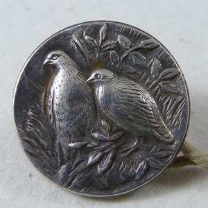 Op de afbeelding: Een zilveren knoop met een reliëfdesign van twee vogels die op een tak met bladeren zitten. De knoop is rond en heeft een glad, glanzend oppervlak.