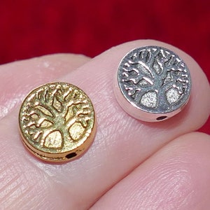 Puede incluir: Dos pequeñas cuentas redondas de metal con un diseño de árbol de la vida. Una cuenta es dorada y la otra plateada. Las cuentas se sostienen entre los dedos sobre un fondo rojo. Cada cuenta tiene un pequeño agujero.