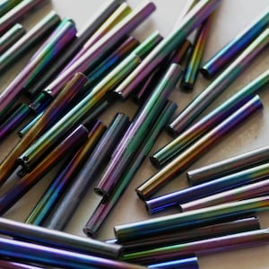 May include: A collection of iridescent glass beads in shades of blue, purple, and green. The beads are long and thin, resembling small rods.