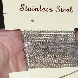 May include: A close-up of a roll of silver stainless steel chain. The chain is on a white card with the words "Stainless Steel" printed in red.
