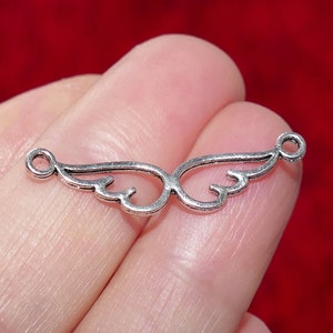 Op de afbeelding: Zilverkleurige metalen bedel met een gedetailleerd ontwerp van twee engelenvleugels. De bedel heeft twee springringen om aan sieraden of andere knutselwerkjes te bevestigen.