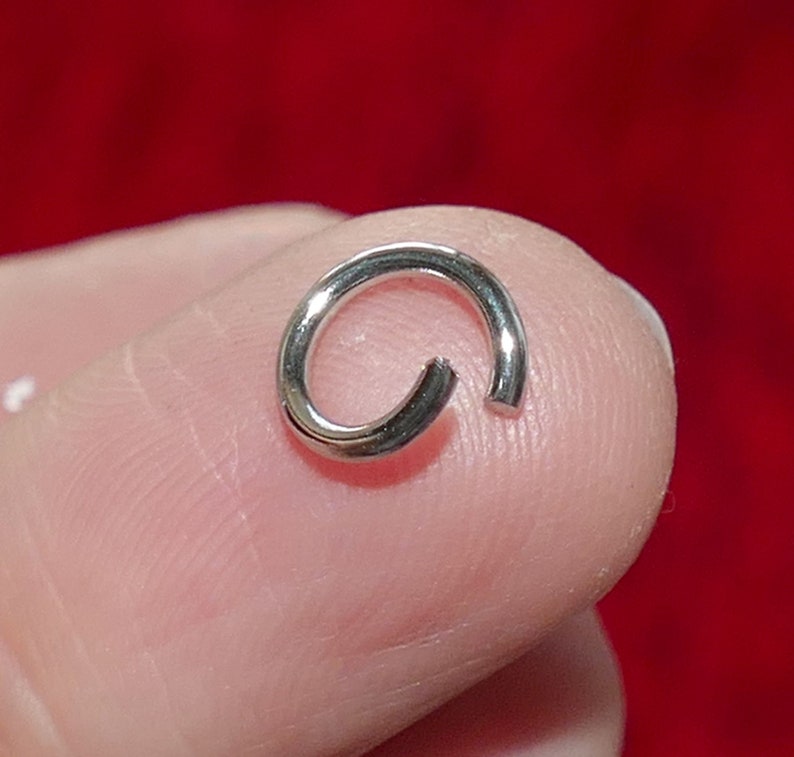 May include: A small, open silver jump ring, likely for jewelry making or crafts. The ring is circular with a small gap, allowing it to be attached to other components. The background is a solid red.