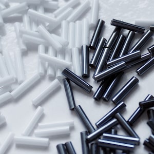 May include: An overhead shot shows a collection of small, cylindrical beads. Some are white, while others are a dark, metallic gray. The beads are scattered across a white surface, creating a contrast between the colors and textures.