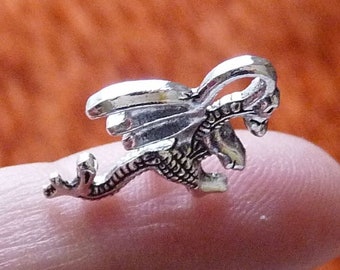 May include: A silver dragon charm with wings outstretched and a detailed design. The charm is on a finger, showing its size.