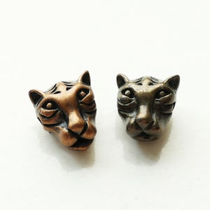 May include: Two metal beads shaped like tiger heads. One bead has a warm copper-toned finish, while the other features a darker, antique bronze-toned finish. Both beads display detailed facial features, including ears, eyes, and nose.