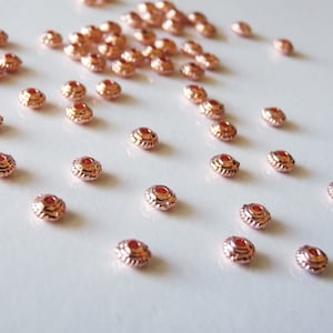 Puede incluir: Una gran cantidad de cuentas de metal de color oro rosa con un pequeño centro rojo. Las cuentas son redondas con un borde elevado.
