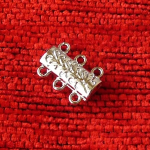 Puede incluir: Conector de cuatro hebras de metal plateado con un diseño floral decorativo. El conector tiene cuatro bucles para sujetar hebras de cuentas u otros componentes de joyería.