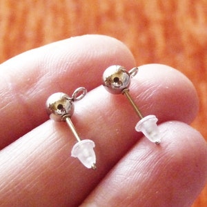 Pode incluir: Dois postes de brincos de prata com tampas de plástico transparente. Os postes têm uma pequena bola na parte superior e um laço para prender os brincos.