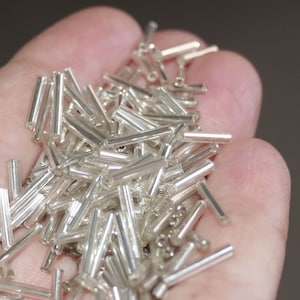 Op de afbeelding: Een grote stapel zilveren glazen kralen in de vorm van lange, dunne cilinders. De kralen zijn verspreid in een hand.