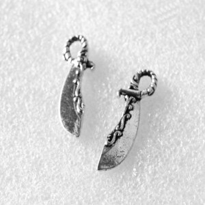 Puede incluir: Dos dijes de metal plateado con forma de espadas curvas o cimitarras. Cada dije presenta un mango texturizado y ornamentado con una anilla en la parte superior para colgar, y una hoja lisa ligeramente afilada con grabados decorativos en el lomo.