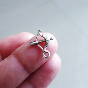 May include: Silver-toned metal charm shaped like a bow and arrow. The charm has a loop at the bottom for attaching to a necklace or bracelet.