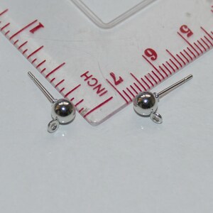 May include: Two silver earring posts with a small round ball at the end and a loop at the bottom. The posts are measured against a ruler.