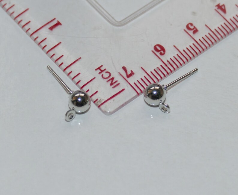May include: Two silver earring posts with a small round ball at the end and a loop at the bottom. The posts are measured against a ruler.