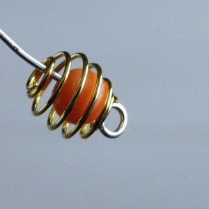 Può includere: Primo piano di una gabbia a spirale color oro con una perla arancione all'interno. La gabbia è attaccata a un filo d'argento. Lo sfondo è grigio chiaro uniforme.