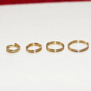 May include: Four gold-colored rings of varying sizes are arranged in a row. Each ring has a double-band design. The rings are displayed on a white surface, with a red background visible at the top.