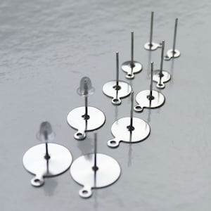 Puede incluir: Un juego de diez postes de pendientes de plata con respaldos redondos. Los postes son de metal y tienen un acabado liso. Los respaldos también son de metal y tienen un acabado ligeramente texturizado.