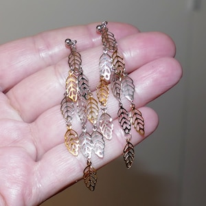 Könnte beinhalten: Hängeohrringe mit Blättermuster. Die Ohrringe haben mehrere Stränge aus durchbrochenen Blättern in Gold- und Silbertönen. Die Ohrringe werden in einer Hand gehalten.