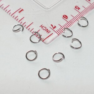 May include: A close-up of eight silver jump rings, scattered on a white surface. The rings are all the same size and are arranged in a random pattern.