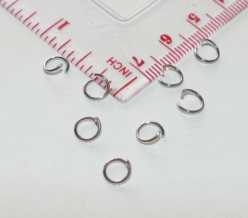 May include: A close-up of eight silver jump rings, scattered on a white surface. The rings are all the same size and are arranged in a random pattern.