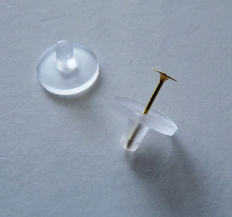 May include: Two clear plastic earring backs, one with a gold post and a flat top, the other with a round top.