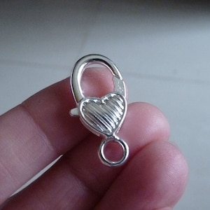 Könnte beinhalten: Silberfarbener herzförmiger Verschluss mit einem Ring zum Anbringen von Schmuck oder anderen Gegenständen.