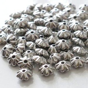 Peut inclure: Un tas de capsules de perles en métal argenté avec un bord festonné. Les capsules de perles sont petites et ont un trou au centre.