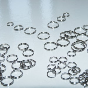 May include: A large quantity of silver key rings, all of different sizes, are scattered on a white surface.