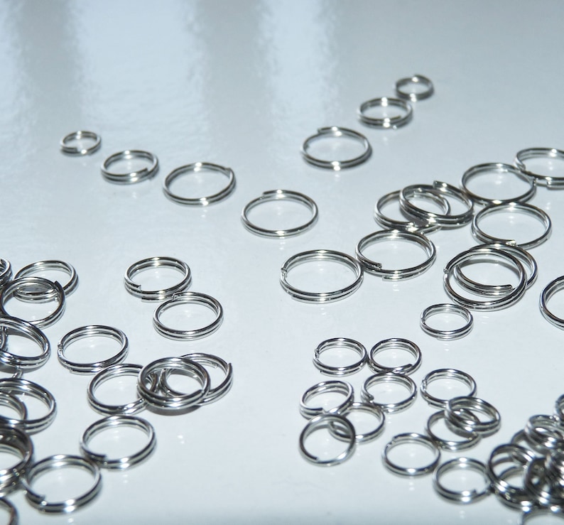 May include: A large quantity of silver key rings, all of different sizes, are scattered on a white surface.
