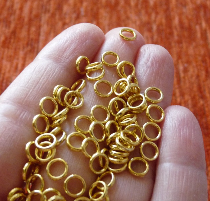 Puede incluir: Una mano sosteniendo una gran cantidad de peque&ntilde;os anillos de salto dorados. Los anillos son todos del mismo tama&ntilde;o y est&aacute;n dispuestos en un patr&oacute;n aleatorio.
