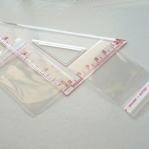 May include: Three clear plastic bags, two of which are partially filled with a clear plastic ruler. The ruler has red markings and the text "INNER TO EXPOSE ADHESIVE" and "INCH" printed on it. The third bag has the text "OKER BRAND" printed on it.