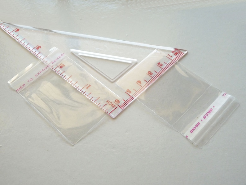 May include: Three clear plastic bags, two of which are partially filled with a clear plastic ruler. The ruler has red markings and the text "INNER TO EXPOSE ADHESIVE" and "INCH" printed on it. The third bag has the text "OKER BRAND" printed on it.