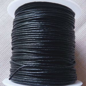 Puede incluir: Una bobina de cordón de algodón encerado negro. El cordón está enrollado firmemente alrededor de una bobina de plástico blanco.