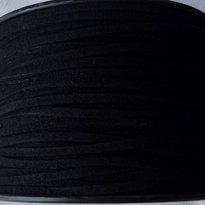 May include: A spool of black suede cord, tightly wound. The cord appears to be flat and approximately 1/8 inch wide. The spool has a black plastic core and is ready for use in crafting or jewelry making.