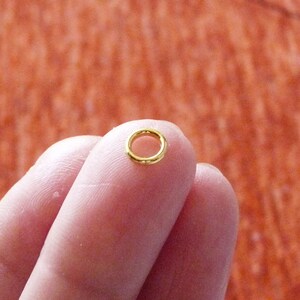 Puede incluir: Un peque&ntilde;o anillo de salto de color dorado, un tipo de hallazgo de joyer&iacute;a, se sostiene entre dos dedos.