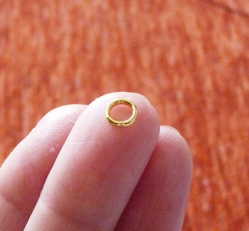 Puede incluir: Un peque&ntilde;o anillo de salto de color dorado, un tipo de hallazgo de joyer&iacute;a, se sostiene entre dos dedos.