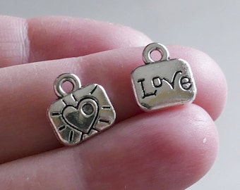 Puede incluir: Dos colgantes de plata. Un colgante presenta un corazón con rayos de sol a su alrededor. El otro colgante es rectangular y dice "Love" en una fuente de escritura.